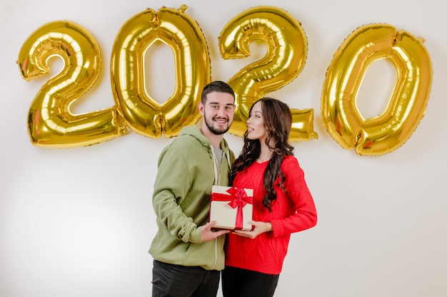 Bonito casal homem e mulher com presente de natal na frente de 2020 balões de ano novo