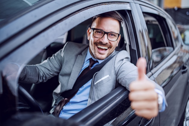 Bonito caucasiano com barba elegante empresário no terno e com óculos, segurando a mão no volante e dando os polegares.