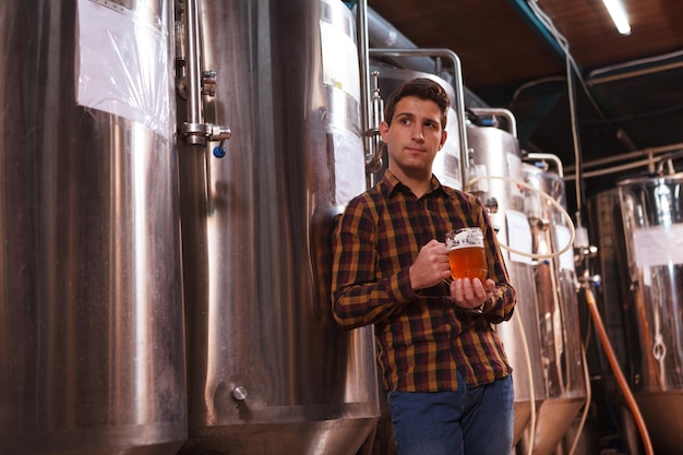 Bonito cervejeiro masculino descansando em sua cervejaria após o dia de trabalho, tomando um copo de cerveja