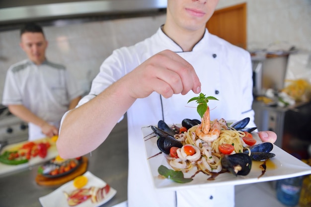 Bonito chef vestido de uniforme branco decorando salada de macarrão e frutos do mar na cozinha moderna