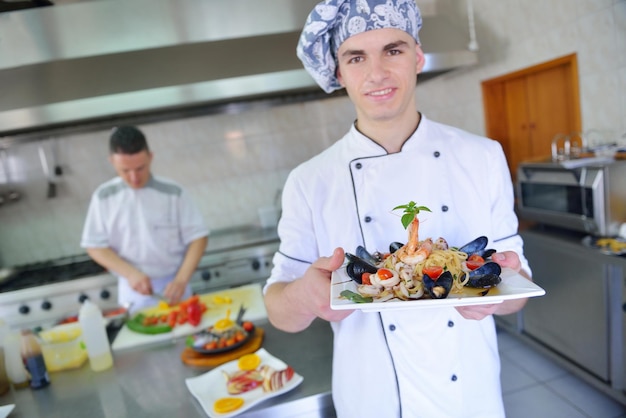 Bonito chef vestido de uniforme branco decorando salada de macarrão e frutos do mar na cozinha moderna