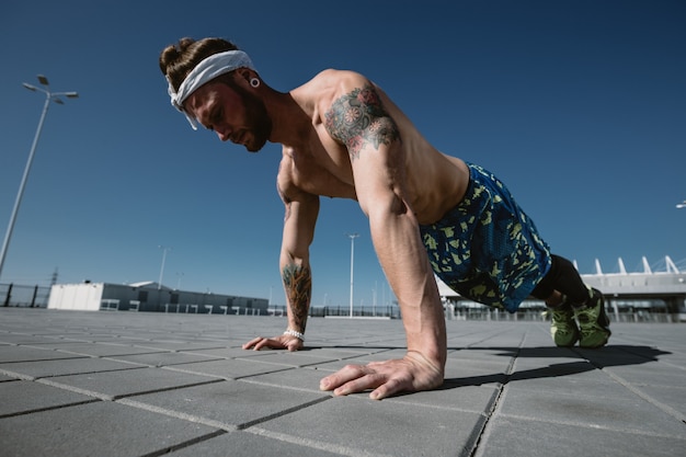 Bonito desportista sem camisa, olhando para longe, fazendo exercícios de flexão no chão de treino ao ar livre em um dia ensolarado. Estilo de vida do esporte.