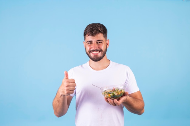 Foto bonito e alegre jovem barbudo vestindo camiseta em pé isolado sobre fundo azul, segurando a tigela com salada fresca polegar para cima