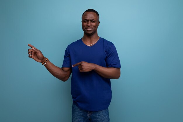 Bonito e amigável jovem africano em uma camiseta azul mostra sua mão em direção ao espaço vazio