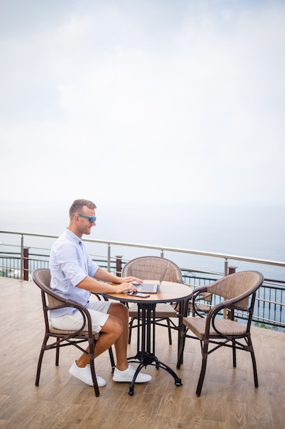 Bonito e bem sucedido jovem empresário sentado à mesa à beira da piscina com um laptop com vista para o mar mediterrâneo. trabalho remoto de férias. conceito de férias