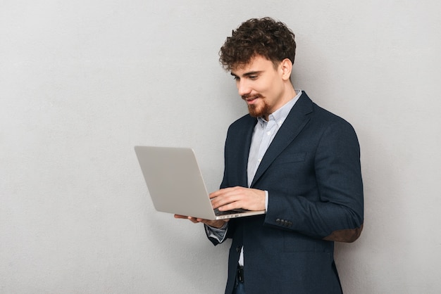 Bonito e confiante jovem empresário vestindo uma jaqueta isolada sobre o cinza, trabalhando em um laptop