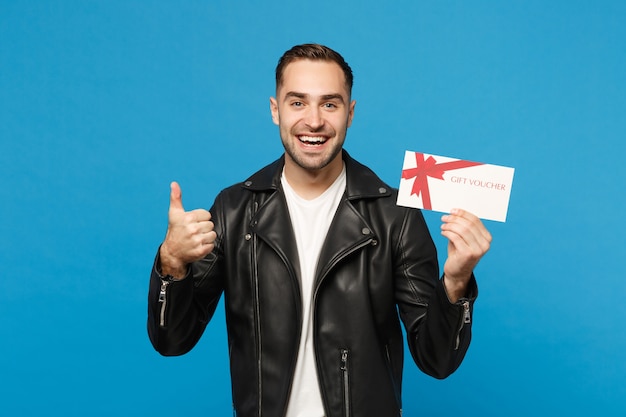 Bonito e elegante jovem com a barba por fazer em t-shirt branca de jaqueta preta segurar certificado de presente isolado no retrato de estúdio de fundo de parede azul. Conceito de estilo de vida de emoções sinceras de pessoas. Simule o espaço da cópia.