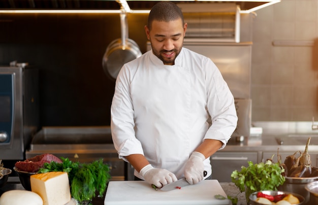 Bonito e jovem chef africano em pé na cozinha profissional em um restaurante que prepara uma refeição de carne e queijo vegetais. retrato de homem em uniforme de cozinheiro corta pimenta vermelha com uma faca de metal.