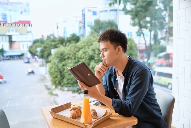 Bonito e jovem designer escreve notas gráficas criativas e fazendo esboço no caderno enquanto está sentado à mesa de madeira no interior do café. homem atencioso escrevendo novas ideias de sucesso para o artigo do blog