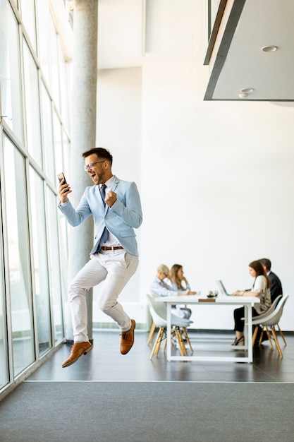 Bonito e jovem empresário com smartphone em um escritório moderno pulando após receber uma ótima notícia