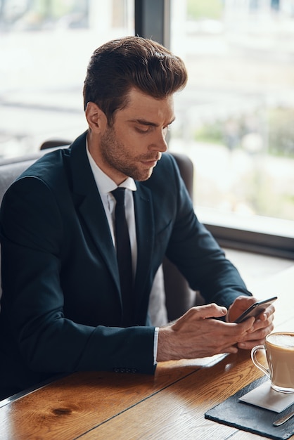 Bonito e jovem empresário em um terno completo usando um telefone inteligente enquanto está sentado no restaurante