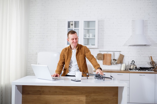 Bonito e jovem empresário trabalhando remotamente com o computador, sentado à mesa da cozinha. Homem bom e feliz sentado na sala segurando um drone nas mãos