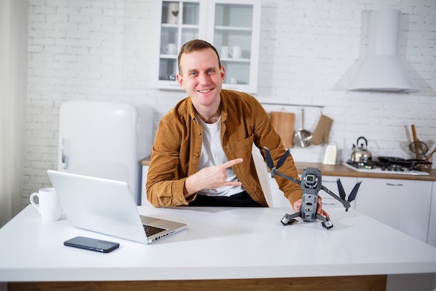 Bonito e jovem empresário trabalhando remotamente com o computador, sentado à mesa da cozinha. Homem bom e feliz sentado na sala segurando um drone nas mãos