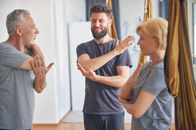 Bonito e jovem treinador branco qualificado, instruindo seus clientes durante os alongamentos de ombro em uma academia