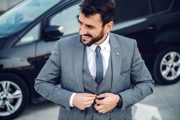 Bonito e sorridente empresário barbudo caucasiano em pé ao ar livre e abotoando o colete. No fundo está o carro dele.