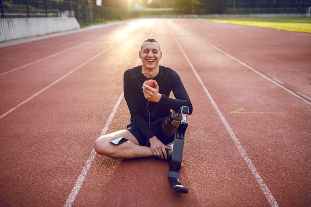 Bonito e sorridente jovem deficiente desportivo caucasiano no sportswear e com uma perna artificial sentado na pista de corrida, ouvindo música e comendo maçã.