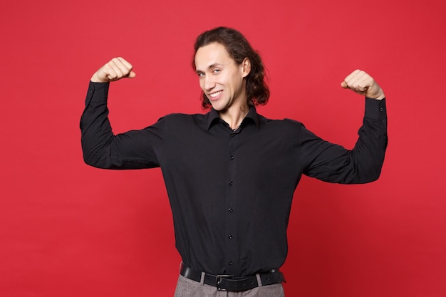 Bonito elegante jovem de cabelos compridos encaracolado na camisa preta posando isolado no retrato de estúdio de fundo vermelho. conceito de estilo de vida de emoções sinceras de pessoas. mock up espaço de cópia. mostrando bíceps, músculos.