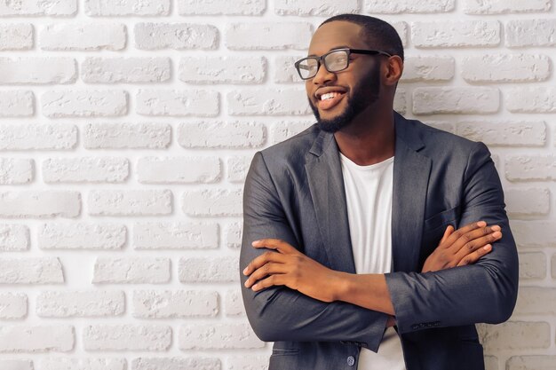Bonito empresário afro-americano na jaqueta clássica cinza e óculos está sorrindo em pé com os braços cruzados contra a parede de tijolos