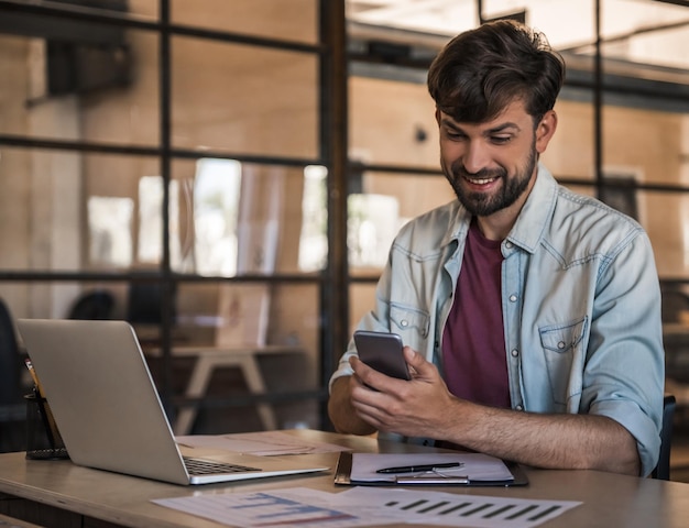 Bonito empresário barbudo em roupas casuais está usando um telefone inteligente e sorrindo enquanto trabalha com um laptop no escritório