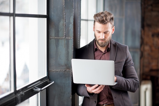 Bonito empresário barbudo vestido de terno em pé com o laptop perto da grande janela no estúdio interior do loft