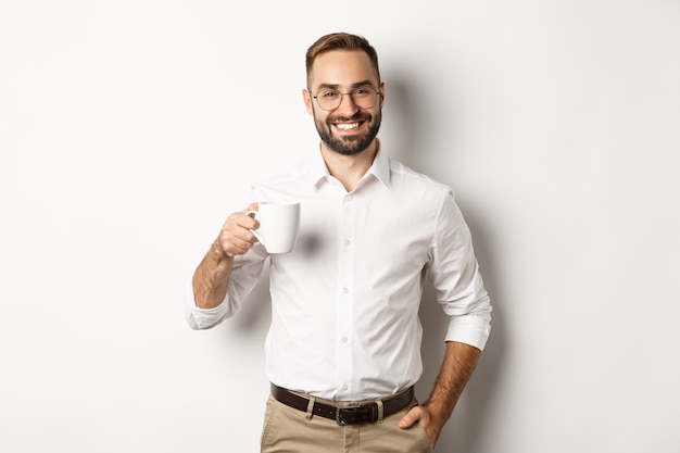 Bonito empresário bebendo café e sorrindo, de pé contra um fundo branco.