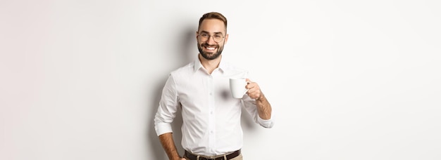 Bonito empresário bebendo café e sorrindo em pé contra um fundo branco