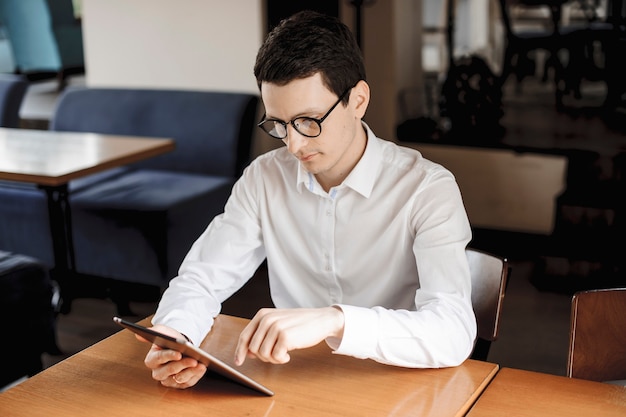 Bonito empresário caucasiano operando em um tablet enquanto está sentado em uma mesa, usando óculos e vestido com uma camisa branca.