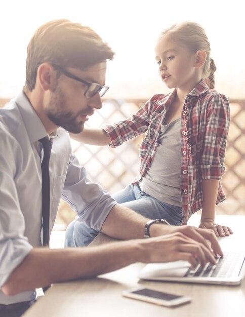 Bonito empresário de óculos está usando um laptop enquanto trabalha em casa sua filha bonitinha está falando com ele