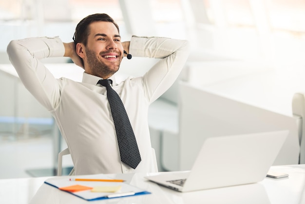 Bonito empresário de terno e fone de ouvido está sorrindo enquanto trabalha com laptop no escritório