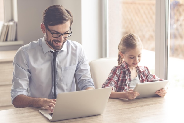 Bonito empresário e sua filha bonitinha estão usando gadgets e sorrindo enquanto passam tempo juntos