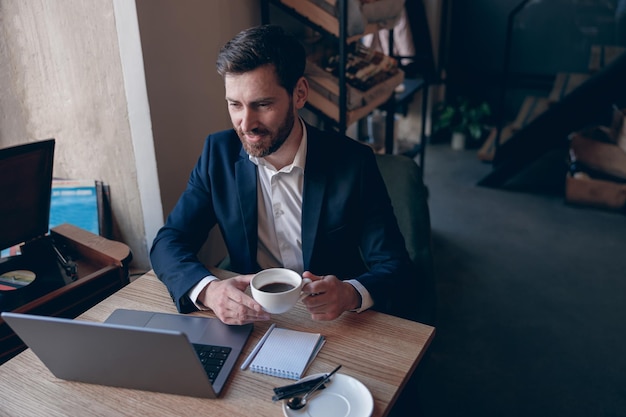 Bonito empresário elegante tomando café e olhando para a tela do laptop no café aconchegante