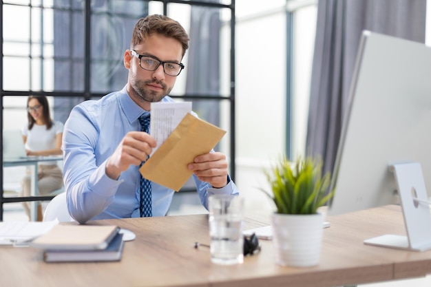 Bonito empresário lendo uma carta do envelope em um desktop no escritório com colegas no fundo