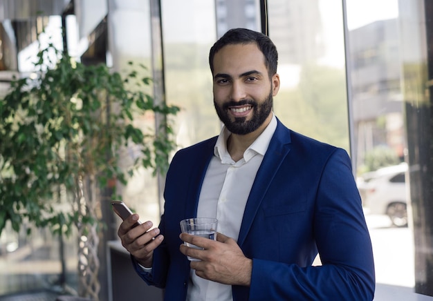 Bonito empresário marroquino sorridente segurando copo de água usando telefone celular no escritório moderno