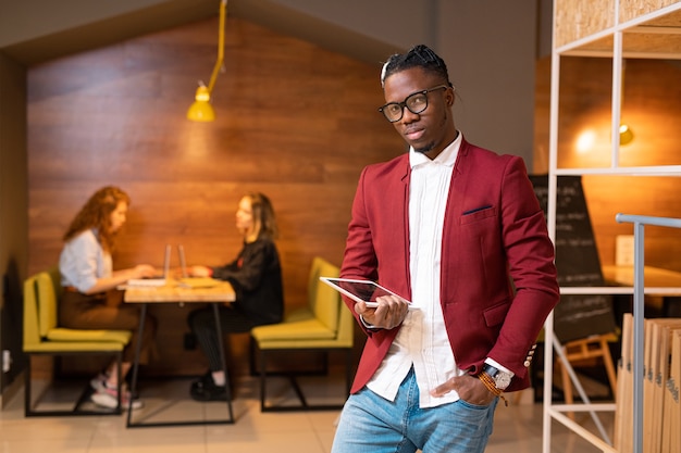 Bonito estudante africano ou empresário contemporâneo com rede touchpad em um café com duas meninas usando laptops