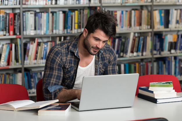 Bonito estudante masculino com laptop e livros trabalhando em uma biblioteca do ensino médio