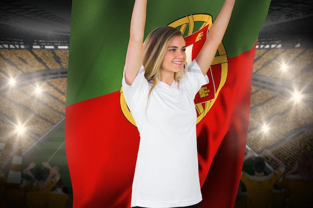 Bonito fanático del fútbol en blanco animando sosteniendo la bandera de portugal contra el vasto estadio de fútbol con fanáticos en amarillo