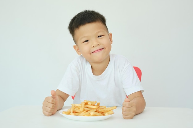 Bonito feliz sorridente menino asiático de 5 anos do jardim de infância comendo batatas fritas batatas fritas sentado à mesa Isolado sobre o fundo da parede branca Conceito de alimentos insalubres para crianças