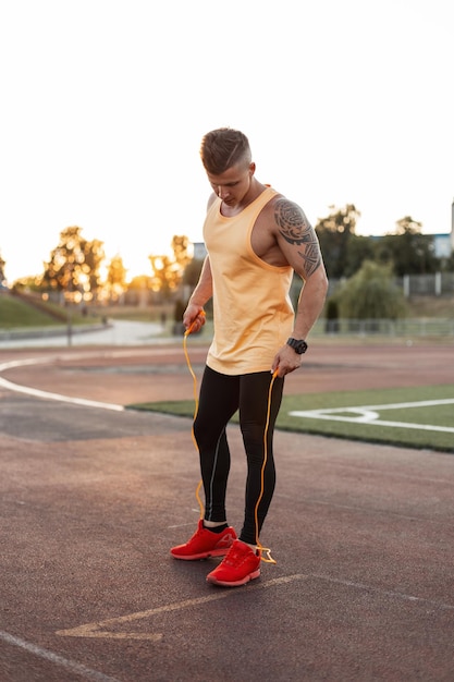 Bonito fitness jovem atleta masculino em roupas esportivas da moda com tênis vermelho com uma corda de pular no estádio Cara atlético se preparando para exercício e treino ao pôr do sol