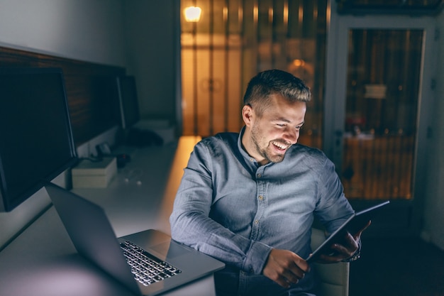 Bonito freelancer barbudo caucasiano com sorriso sentado no escritório tarde da noite e usando o tablet.