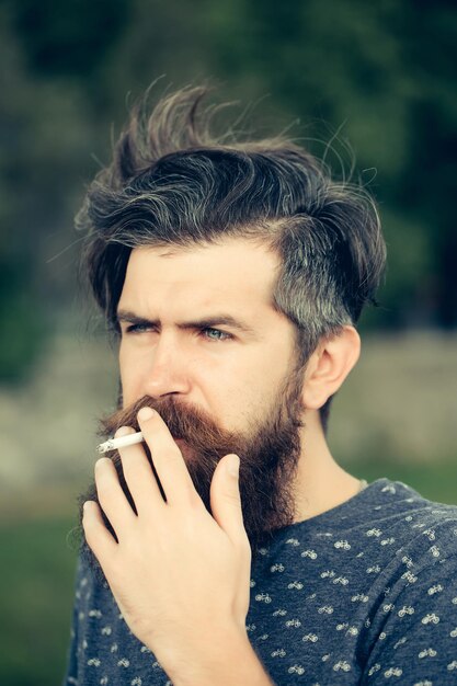 Bonito hipster barbudo sexy jovem com barba longa e bigode tem cabelo elegante na cara peluda séria fumando cigarro ao ar livre no fundo verde natural closeup