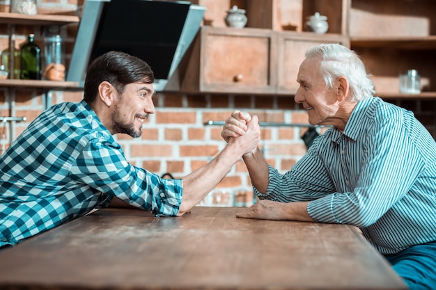 Bonito hombre positivo feliz sosteniendo la mano de su padre y sonriendo mientras hace pulseadas