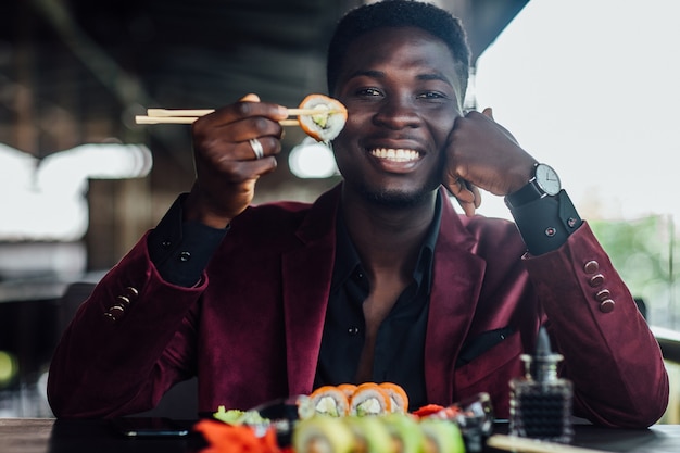 Bonito homem africano com prato de sushi na esplanada. Homem de negócios feliz prova sushi.