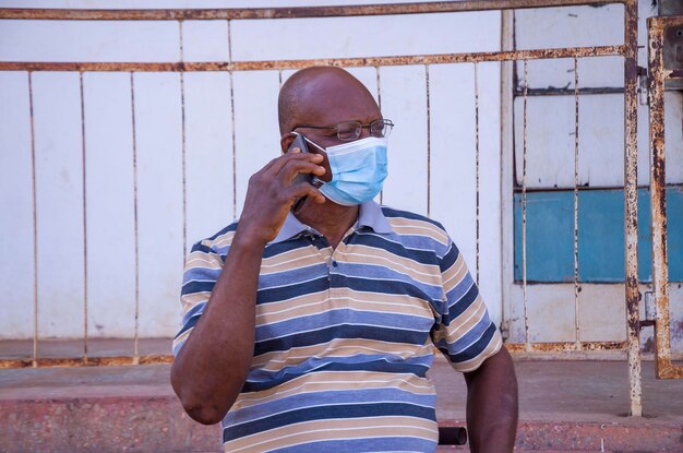 Bonito homem africano idoso usando máscara facial prevenindo, evitou o surto na sociedade se sentindo animado com a ligação que está fazendo com seu celular.