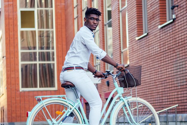 Bonito homem afro-americano elegante bem vestido em pé com um fundo de construção de tijolo vermelho de bicicleta azul. conceito de transporte ecológico esportivo.