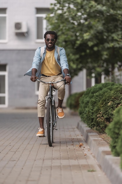 Foto bonito homem afro-americano elegante sorrindo enquanto andava de bicicleta ao longo da rua