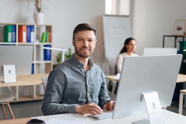 Bonito homem alegre sentado na mesa usando computador moderno no escritório de espaço aberto