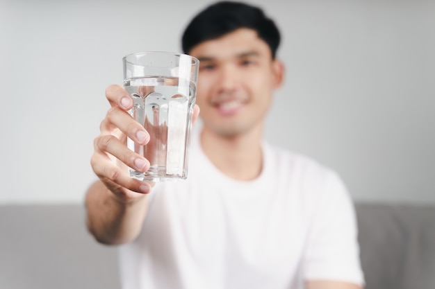 Bonito homem asiático bebendo um copo d'água no sofá da sala