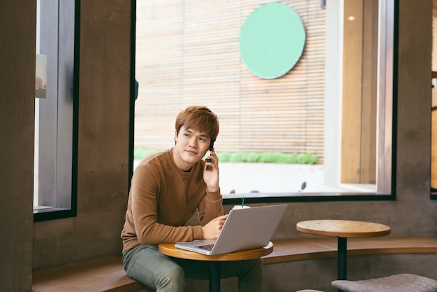 Bonito homem asiático usando laptop sentado na mesa de madeira em uma cafeteria com uma xícara de café