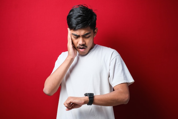 Bonito homem asiático vestindo camiseta branca olhando para o relógio preocupado, com medo de se atrasar por causa do fundo vermelho