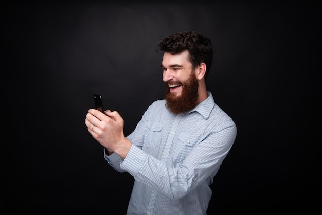Bonito homem barbudo alegre usando móveis, digitando sms, em cima de fundo escuro isolado